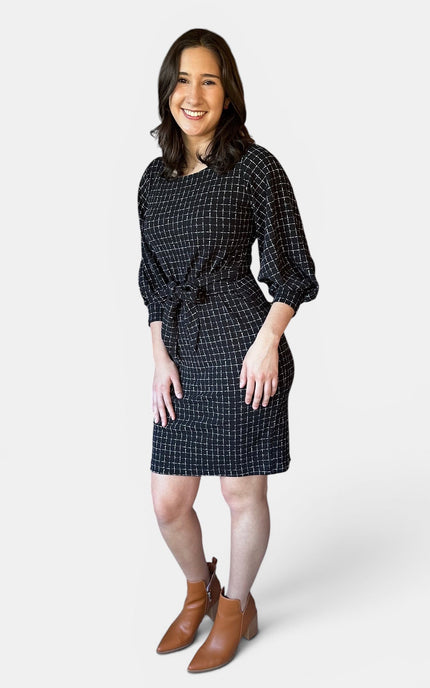 A woman modeling a black checkered mini dress with three-quarter sleeves and a tie waist, paired with brown ankle boots, against a light gray studio background.
