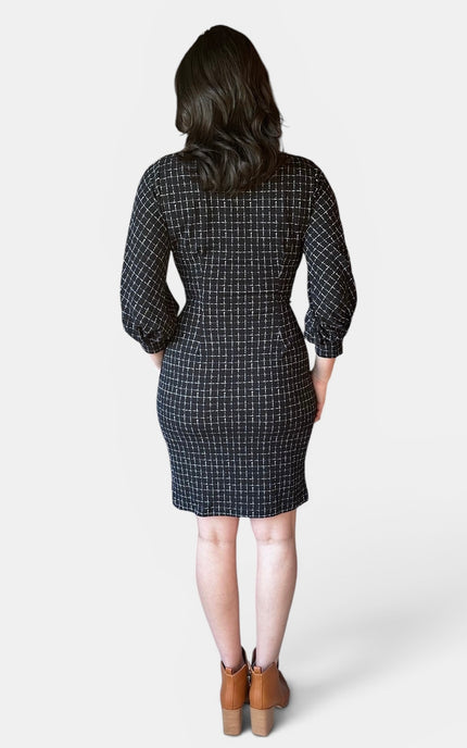 A woman viewed from the back wearing a black grid-patterned knee-length dress with three-quarter sleeves, paired with brown ankle boots, against a light gray studio background.
