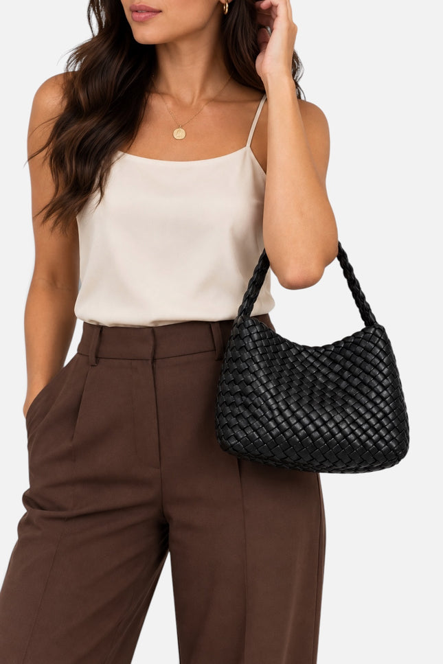 A woman wearing a cream camisole and brown trousers carries a black woven leather shoulder bag against a light gray studio background.