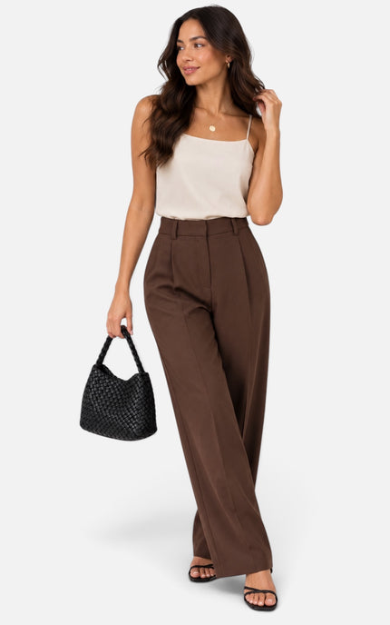 A woman modeling high-waisted, wide-leg brown trousers paired with a cream camisole top, black sandals, and a black woven handbag, set against a plain light gray studio background.