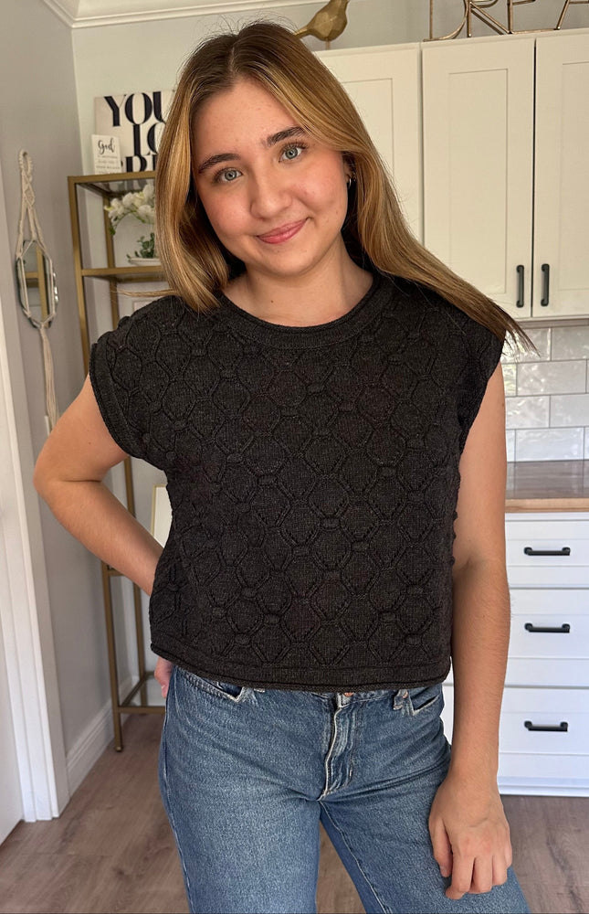 Woman wearing a black sleeveless knitted top and blue jeans in a kitchen.