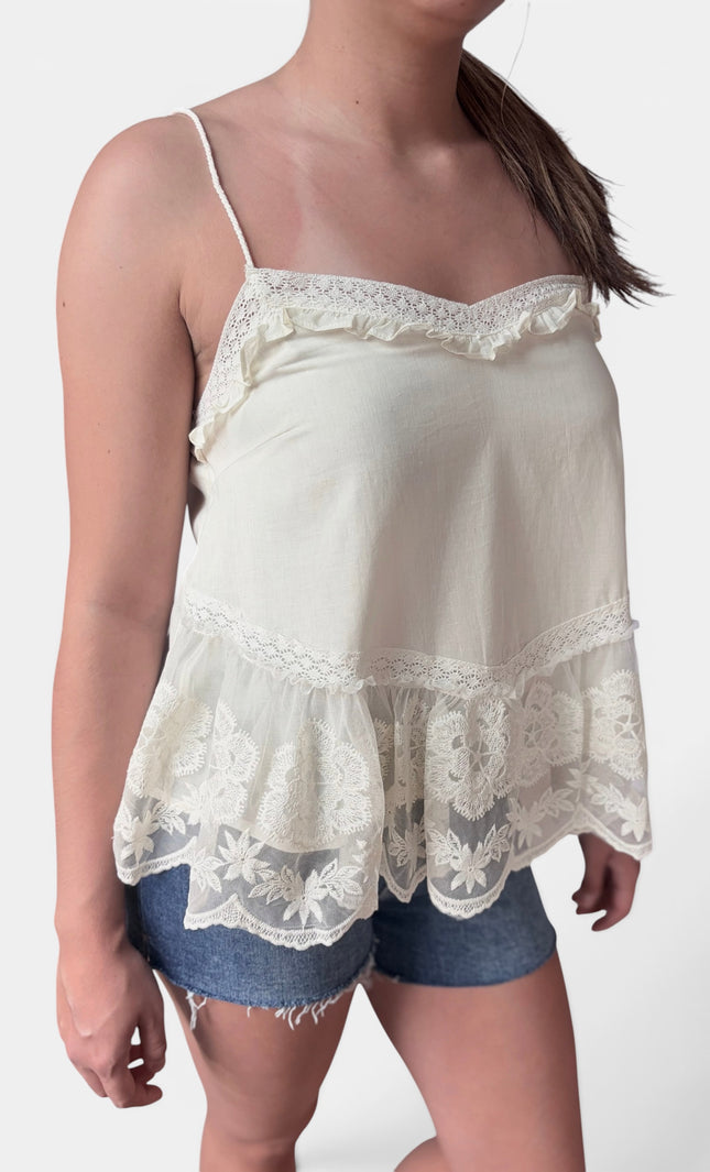 A woman wearing a cream-colored camisole top with delicate lace trim and ruffles, paired with denim shorts, against a plain light background.