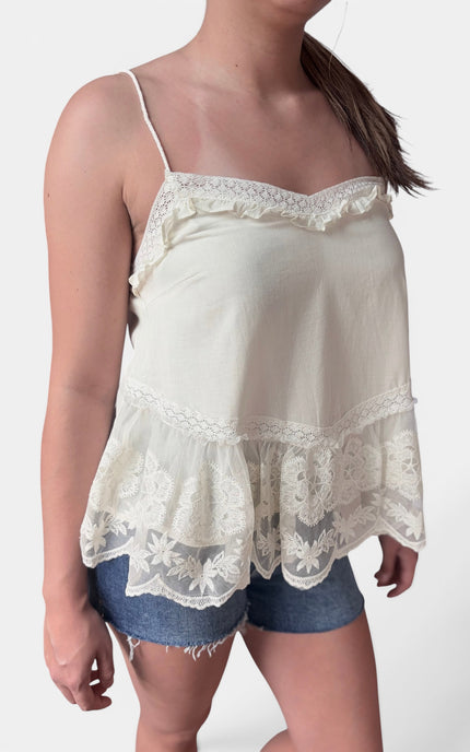A woman wearing a cream-colored camisole top with delicate lace trim and ruffles, paired with denim shorts, against a plain light background.