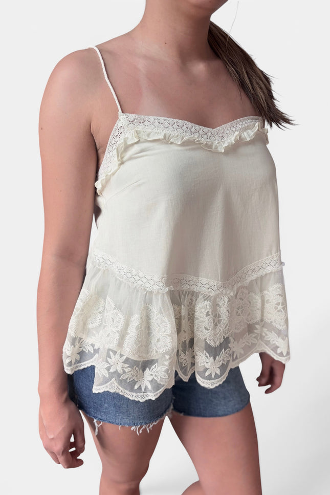 A woman wearing a cream-colored camisole top with delicate lace trim and ruffles, paired with denim shorts, against a plain light background.