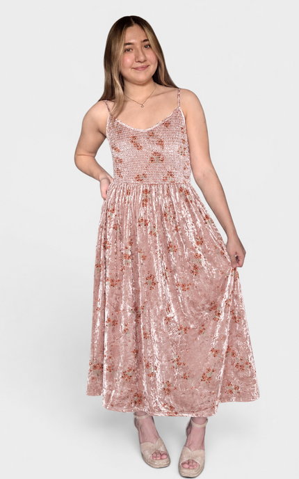 A woman modeling a sleeveless, midi-length pink velvet dress with a subtle floral pattern, photographed against a solid light gray background.