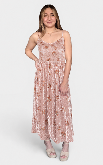 A woman modeling a sleeveless, midi-length pink velvet dress with a subtle floral pattern, photographed against a solid light gray background.