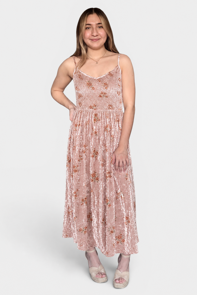 A woman modeling a sleeveless, midi-length pink velvet dress with a subtle floral pattern, photographed against a solid light gray background.