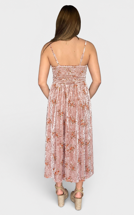 A woman viewed from the back wearing a sleeveless midi dress with thin straps, a smocked bodice, and a floral pattern on a pinkish velvet fabric, paired with beige espadrille heels against a light gray studio background.