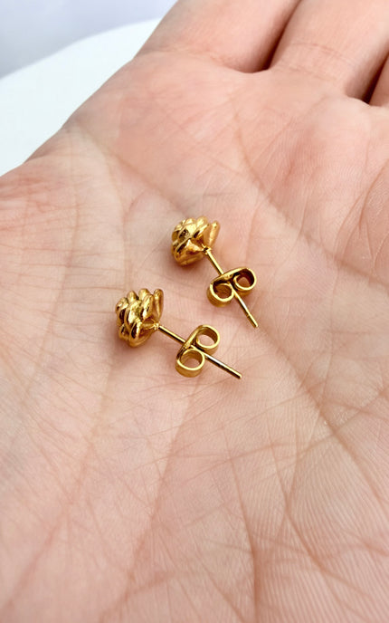 A close-up view of a pair of gold rose stud earrings held gently in the palm of a hand, showcasing the intricate, sculpted petal texture and romantic design.