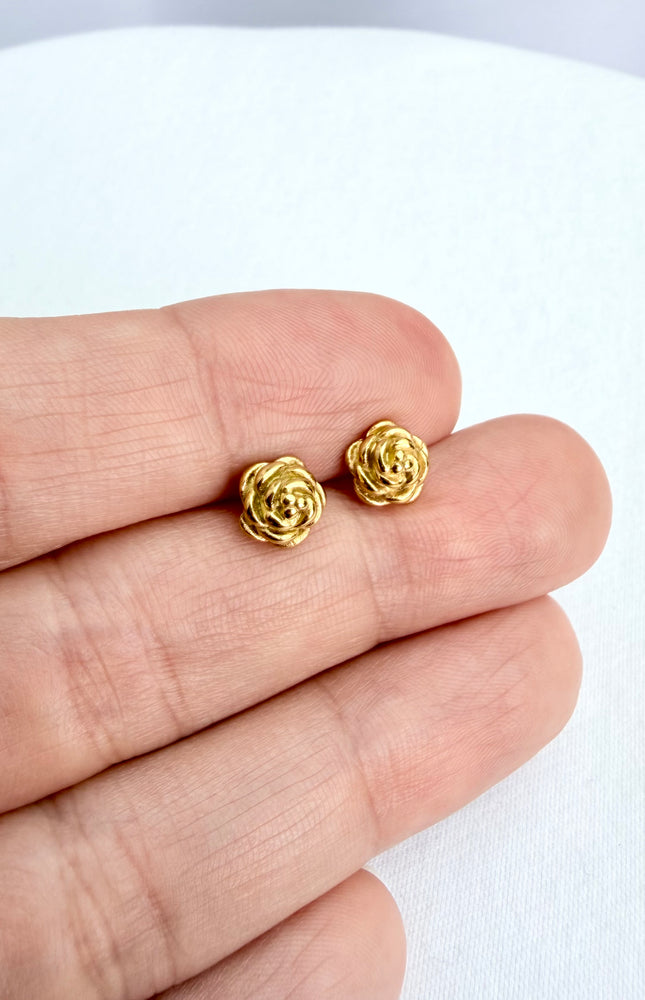 A detailed, high-texture shot of a tiny 18k gold PVD plated rose stud earring resting between fingers, highlighting the light-catching details of the floral design.
