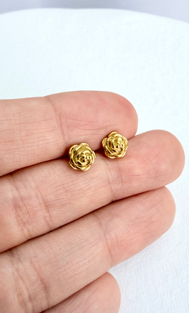A detailed, high-texture shot of a tiny 18k gold PVD plated rose stud earring resting between fingers, highlighting the light-catching details of the floral design.