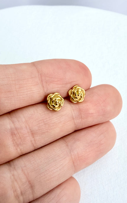 A detailed, high-texture shot of a tiny 18k gold PVD plated rose stud earring resting between fingers, highlighting the light-catching details of the floral design.