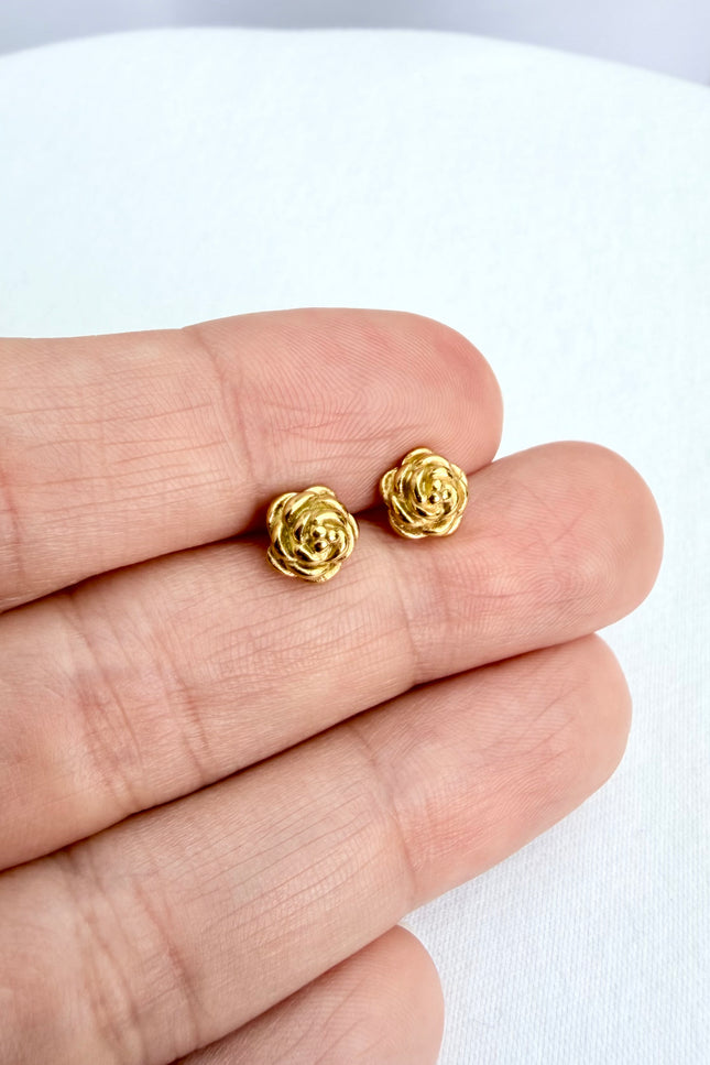 A detailed, high-texture shot of a tiny 18k gold PVD plated rose stud earring resting between fingers, highlighting the light-catching details of the floral design.