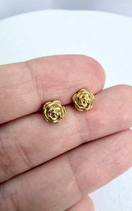 A detailed, high-texture shot of a tiny 18k gold PVD plated rose stud earring resting between fingers, highlighting the light-catching details of the floral design.