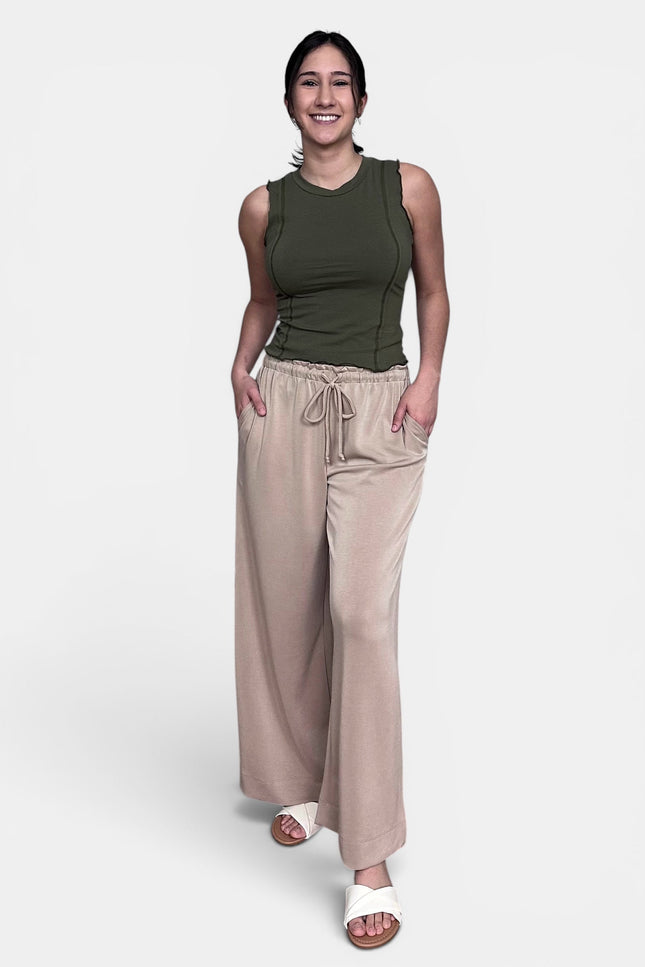 A woman modeling a sleeveless olive green top and wide-leg beige drawstring pants against a light gray studio background.
