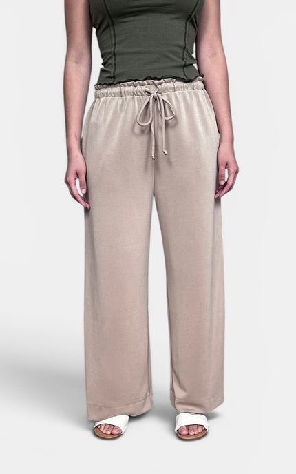 A model wearing beige wide-leg lounge pants with an elastic drawstring waist, paired with an olive green top and white slide sandals, against a light gray studio background.