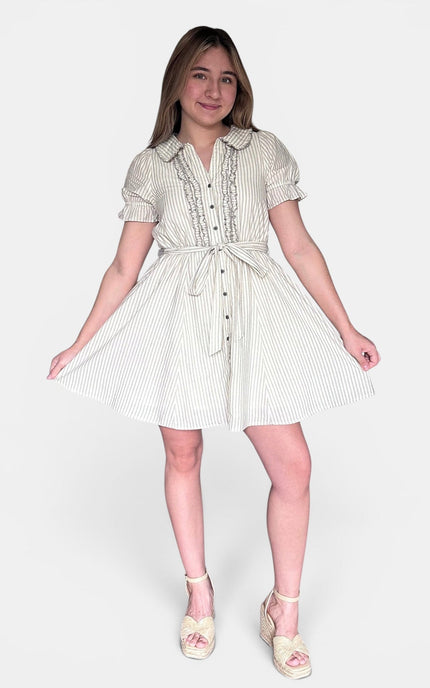 A young girl modeling a short-sleeve, button-front dress with vertical stripes in cream and a light pastel pink, paired with light pink wedge sandals, against a plain light gray background.