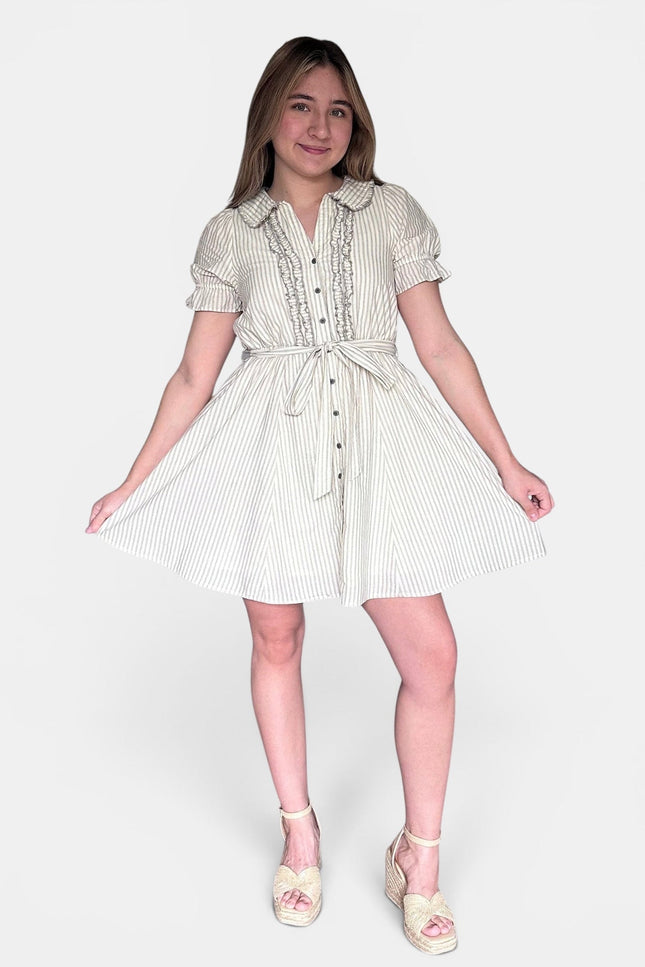A young girl modeling a short-sleeve, button-front dress with vertical stripes in cream and a light pastel pink, paired with light pink wedge sandals, against a plain light gray background.