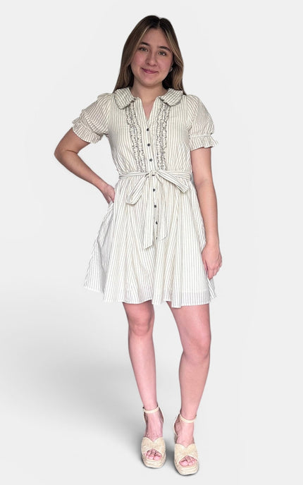 A young woman modeling a short-sleeve cream and white vertically striped shirt dress with ruffle trim, a buttoned front, and a tie waist, paired with cream platform sandals, against a light gray studio background.
