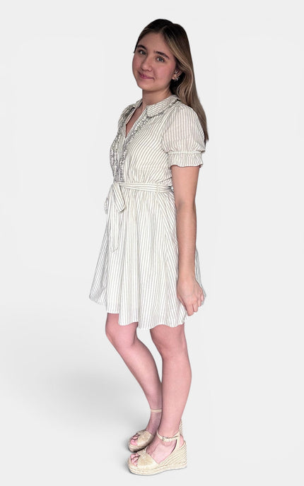 A young woman modeling a short-sleeved cream and white vertically striped dress with a tie waist, standing against a plain light gray studio background.