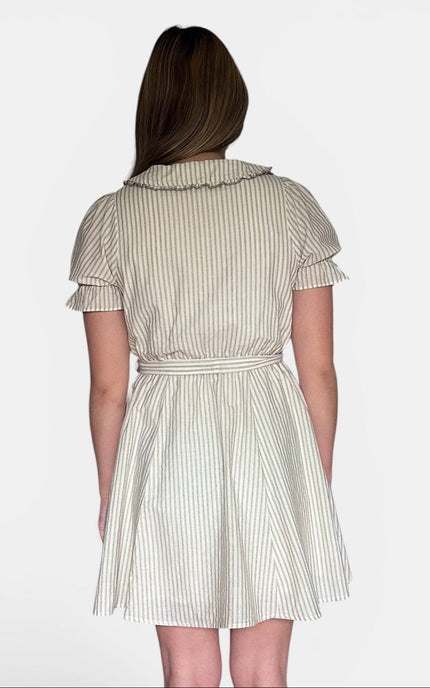 A woman viewed from the back wearing a short-sleeve cream dress with thin vertical green stripes, a ruffled collar, and a tie belt at the waist, set against a plain light gray background.