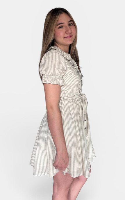 A young woman modeling a short-sleeve cream dress with thin vertical navy stripes, a ruffled collar, and a matching fabric tie belt, shown against a plain light gray background.