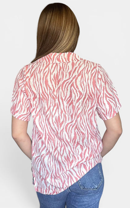 A woman viewed from the back wearing a short-sleeve collared button-up shirt with a pink and white zebra-like abstract print, paired with blue jeans, against a light gray studio background.