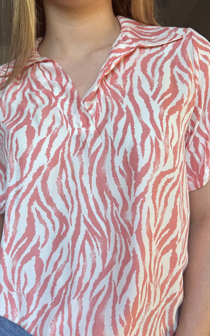 Close up of short-sleeve collared blouse with a coral pink and white zebra-like abstract pattern, paired with blue jeans against a plain light background.