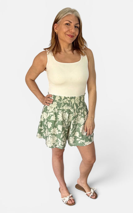 A woman modeling a sleeveless cream top and green floral print shorts against a light gray studio background.