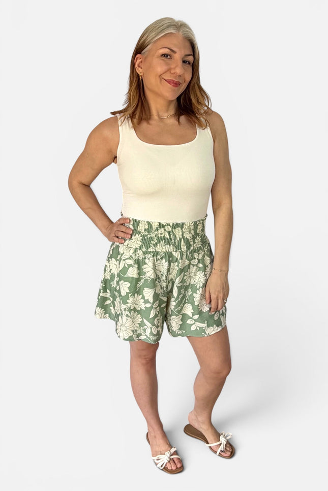A woman modeling a sleeveless cream top and green floral print shorts against a light gray studio background.