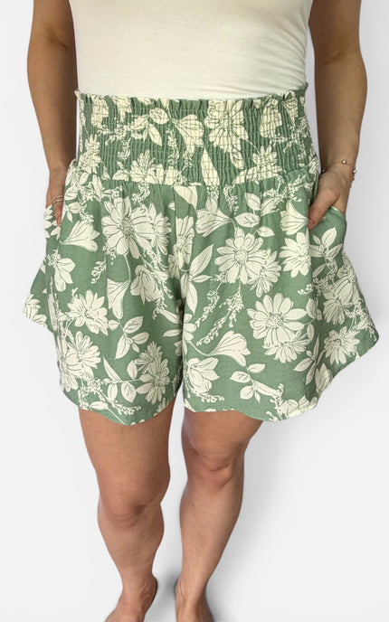 A person wearing green and off-white floral print shorts with a smocked waistband and side pockets, paired with a cream top against a light gray studio background.