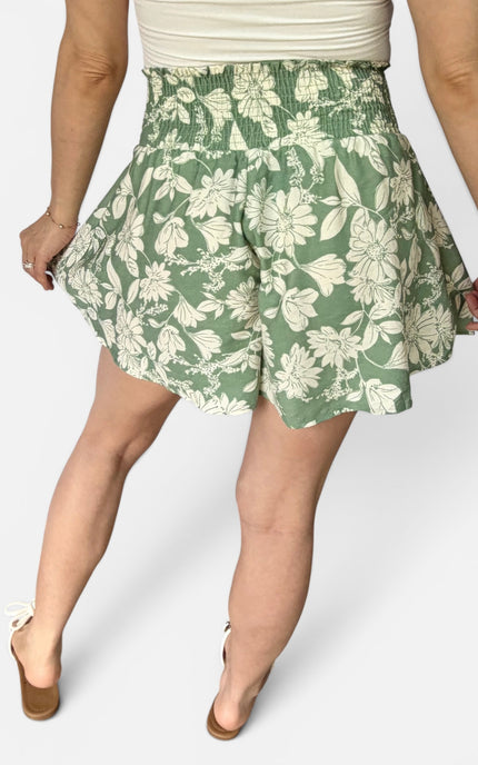 A person wearing a pair of high-waisted, flared shorts with a sage green and cream floral pattern, paired with a cream top and tan wedge sandals, shown from the waist down against a light gray studio background.