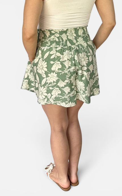 A woman wearing a green and white floral print mini skirt with a smocked waistband, paired with a cream top and white slide sandals, shown from the back against a light gray studio background.