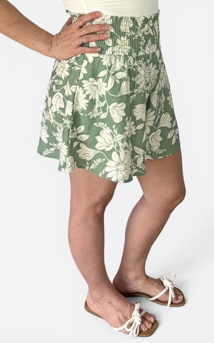 A person wearing a green and white floral print skort with an elastic waistband, paired with white slide sandals, shown from the waist down on a light gray background.