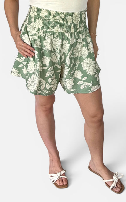 A person wearing green and white floral print shorts, a cream top, and white slide sandals, shown from the waist down against a light gray studio background.