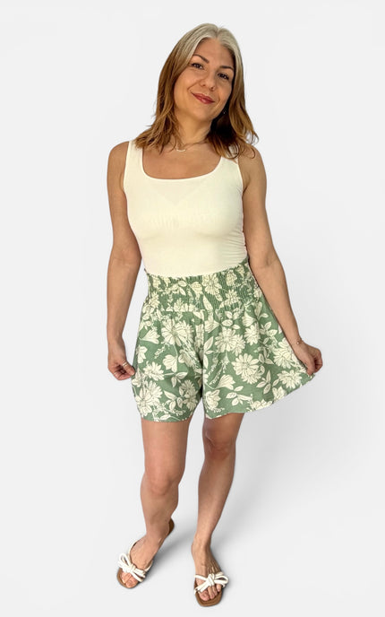 A woman modeling a sleeveless cream top and green floral print shorts against a light gray studio background.