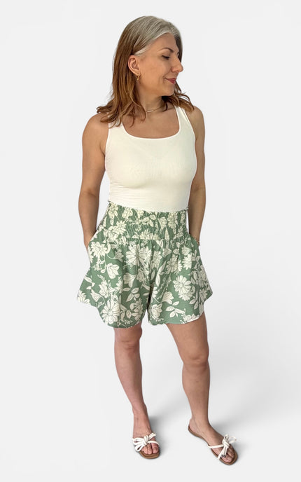 A woman modeling a sleeveless cream tank top and green floral shorts against a light gray studio background.