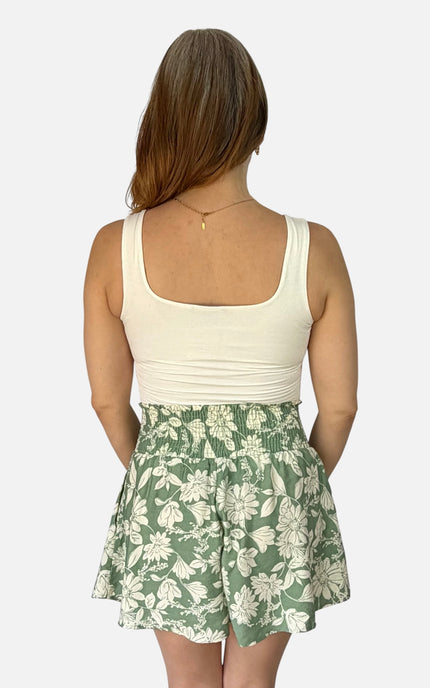 A woman shown from the back wearing a cream scoop-neck tank top and green floral print shorts against a light gray background.