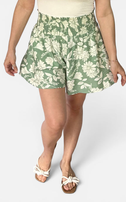 A person wearing green and white floral print shorts, a cream top, and white slide sandals, shown from the waist down against a light gray studio background.