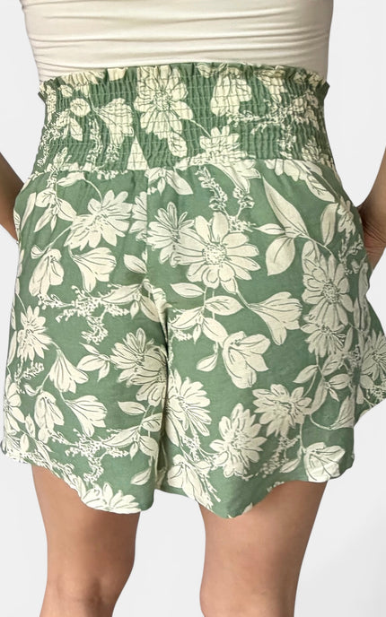 A close-up of a person wearing green smocked shorts with a large white floral print, paired with a white ribbed top.