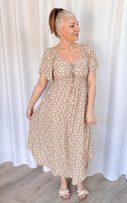 A woman modeling a beige short-sleeve midi dress with a white floral pattern, standing in front of a white curtain backdrop.