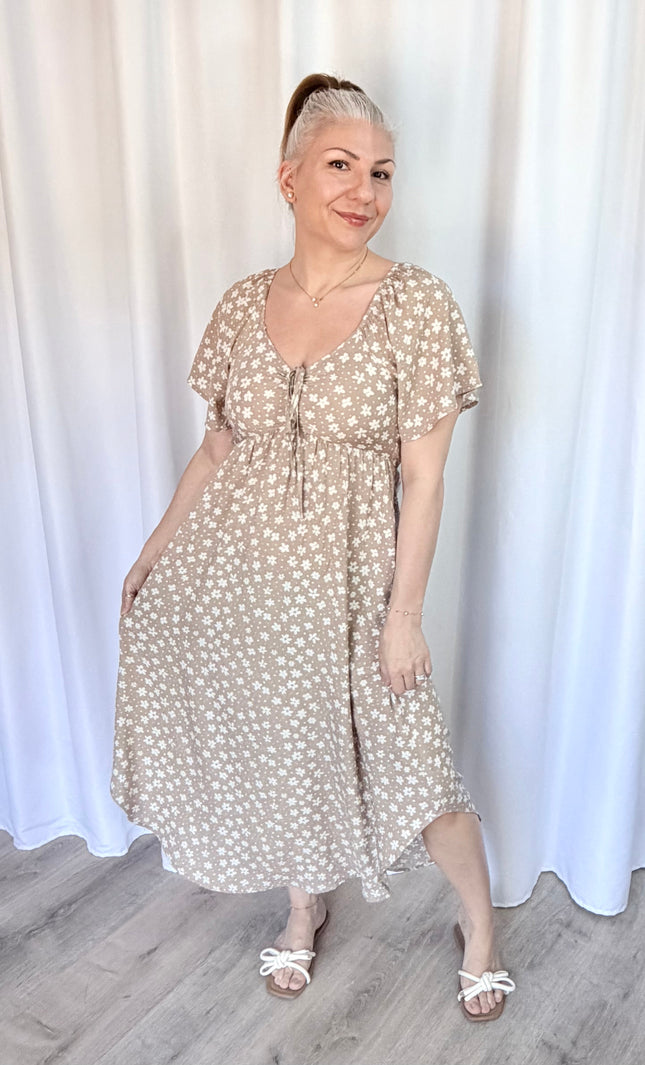 Woman wearing a beige floral dress standing against a white curtain.