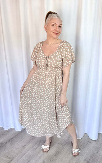 Woman wearing a beige floral dress standing against a white curtain.