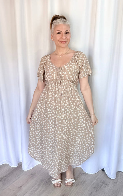 Woman wearing a beige floral dress standing against a white curtain.