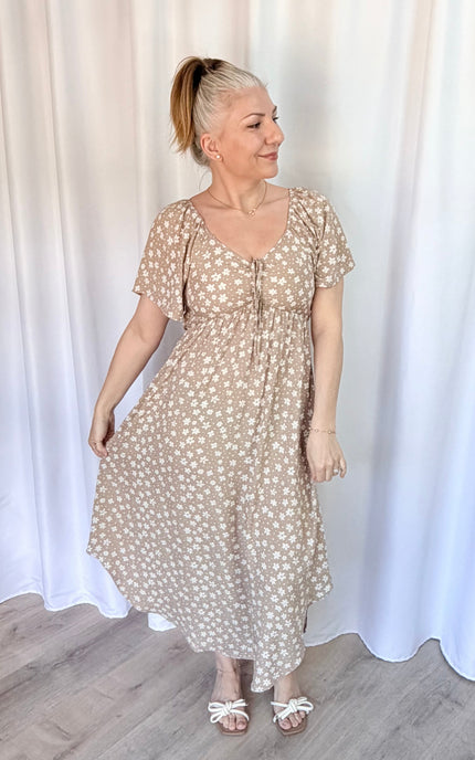 Woman wearing a beige floral dress standing against a white curtain.