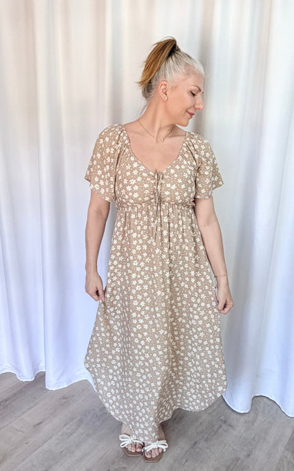 Woman wearing a beige floral dress standing against a white curtain.