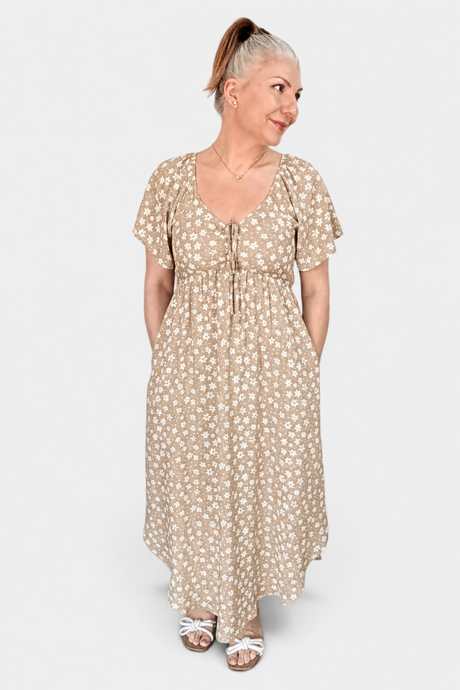 A woman modeling a beige maxi dress with a white ditsy floral print, featuring short flutter sleeves, a V-neckline with tie-front details, and side pockets, set against a plain light gray background.