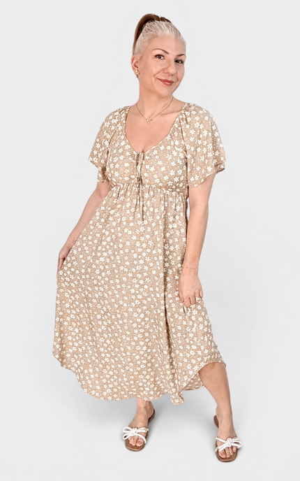 A woman modeling a beige midi dress with a white floral print, featuring short puff sleeves, a V-neckline with a tie-front detail, and a gathered waist. She is wearing white slide sandals against a plain light gray studio background.