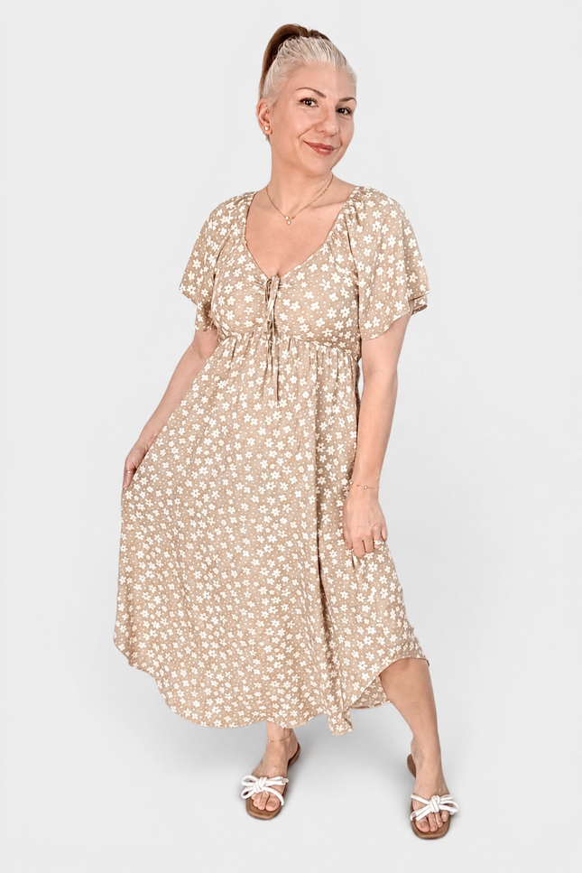 A woman modeling a beige midi dress with a white floral print, featuring short puff sleeves, a V-neckline with a tie-front detail, and a gathered waist. She is wearing white slide sandals against a plain light gray studio background.