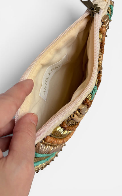 A close-up of a hand holding open a zippered pouch, showing the interior lining and a decorative exterior covered with metallic gold beads, brown shells, and turquoise accents. The brand name 'ANTIK KRAKOW' is visible on the interior tag.